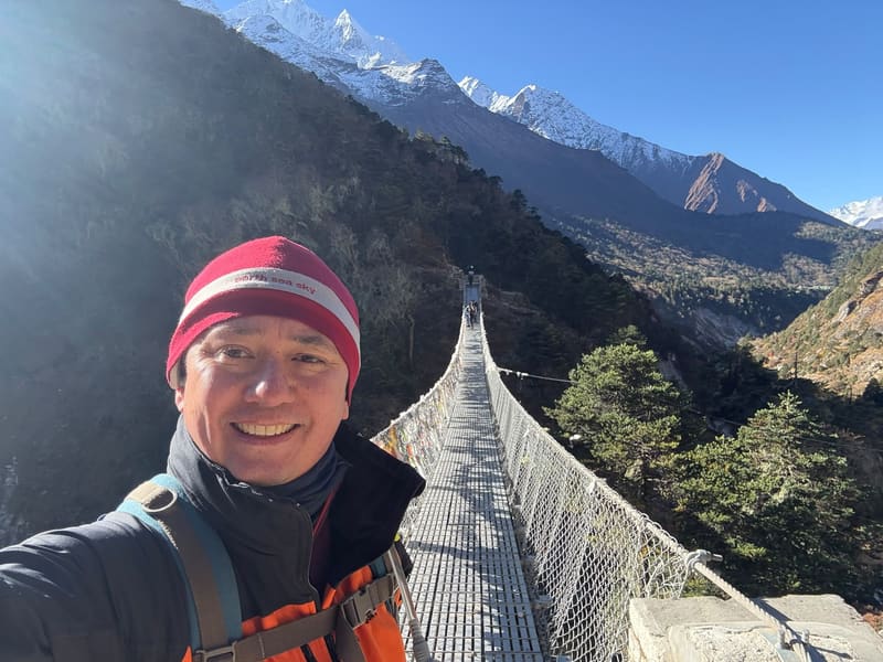 David smiling on a suspension bridge strung with prayer flags, snow-capped peaks behind