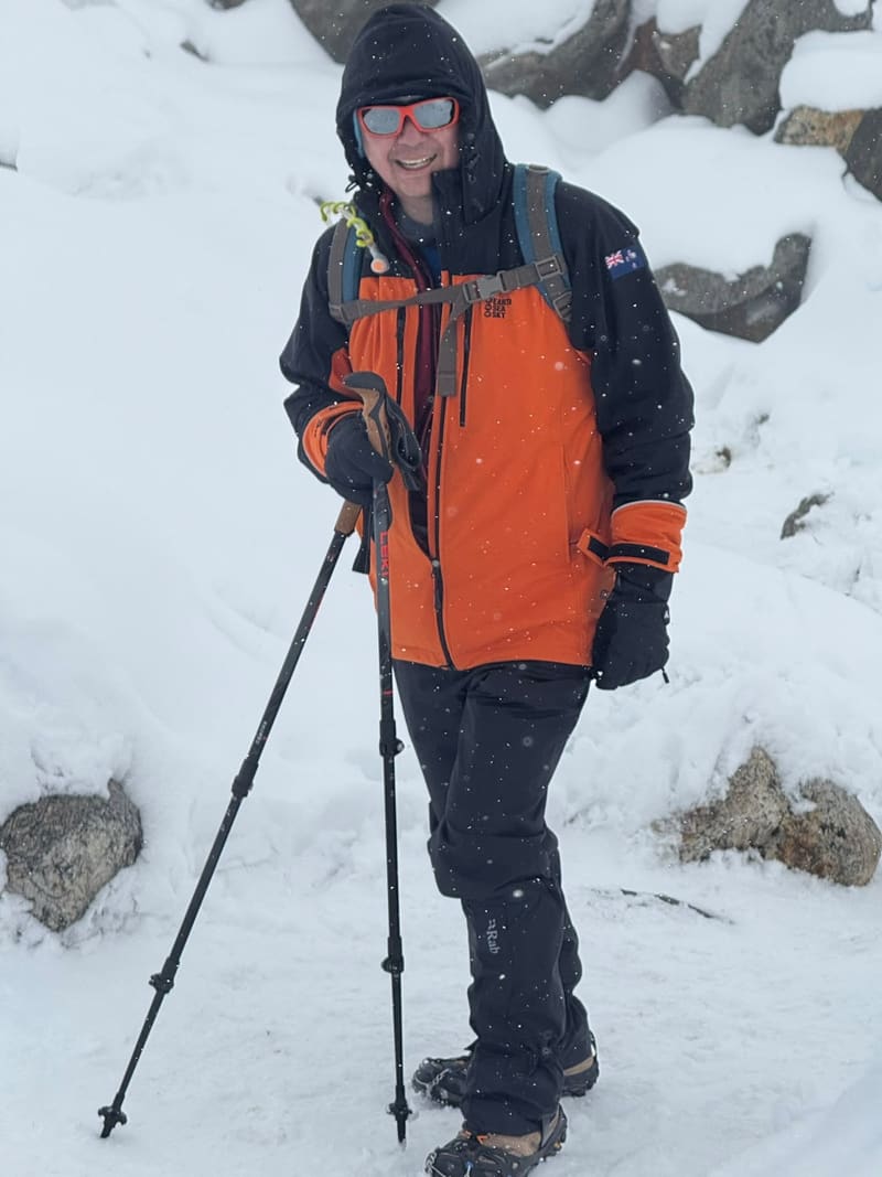 David in an orange jacket with a New Zealand flag patch, smiling in falling snow with trekking poles